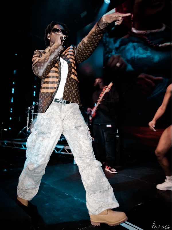 Shallipopi wearing a patterned jacket and light-colored cargo pants singing into a microphone on a dark stage. — concert photography London by Lamss Media